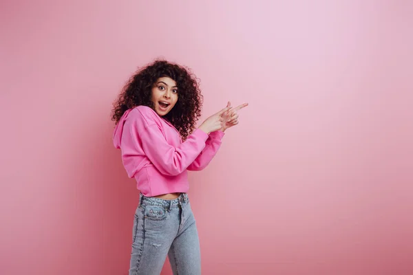 Schockiertes zweirassiges Mädchen, das in die Kamera schaut, während es mit den Fingern auf rosa Hintergrund zeigt — Stockfoto