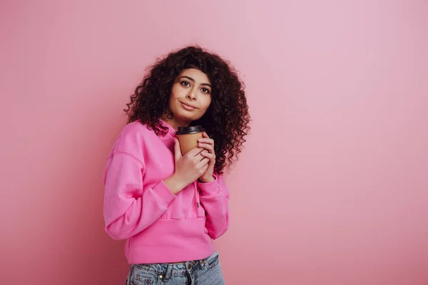 Позитивна, мрійлива бі-расова дівчина дивиться геть, тримаючи каву на рожевому фоні — стокове фото