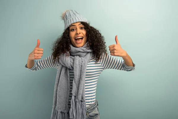 Alegre bi-racial chica en cálido sombrero y bufanda mostrando los pulgares hacia arriba mientras se mira a la cámara sobre fondo gris - foto de stock