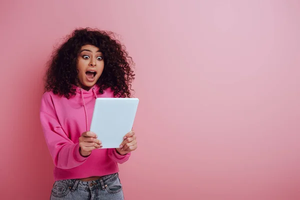 Shocked bi-racial girl looking at digital tablet on pink background — Stock Photo