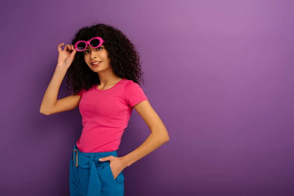 Симпатичная двурасовая девушка касается солнцезащитных очков, улыбаясь в камеру на фиолетовом фоне — стоковое фото