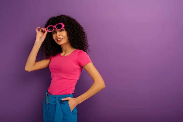 Menina bonita bi-racial tocando óculos de sol enquanto sorrindo para a câmera no fundo roxo — Fotografia de Stock