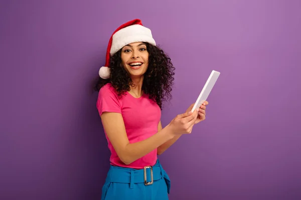 Счастливая девушка смешанной расы в шляпе санта, держащая цифровую табличку, глядя в камеру на фиолетовом фоне — стоковое фото