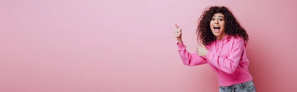 Panoramic shot of excited bi-racial girl showing thumbs up on pink background — Stock Photo