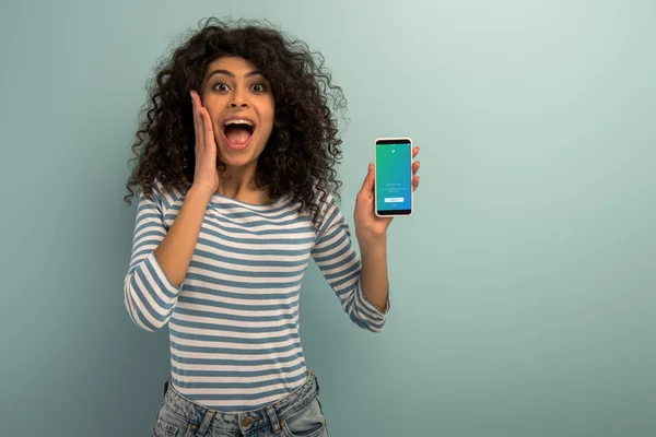 1KYIV, UKRAINE - NOVEMBER 26, 2019: excited bi-racial girl looking at camera while showing smartphone with Twitter app on grey background — Stock Photo