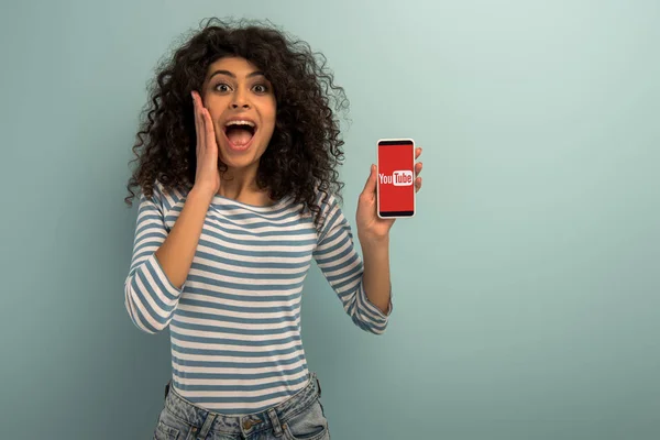 KYIV, UKRAINE - NOVEMBER 26, 2019: excited bi-racial girl looking at camera while showing smartphone with Youtube app on grey background — Stock Photo