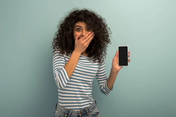 Shocked bi-racial girl covering mouth with hand while showing smartphone with blank screen on grey background — Stock Photo