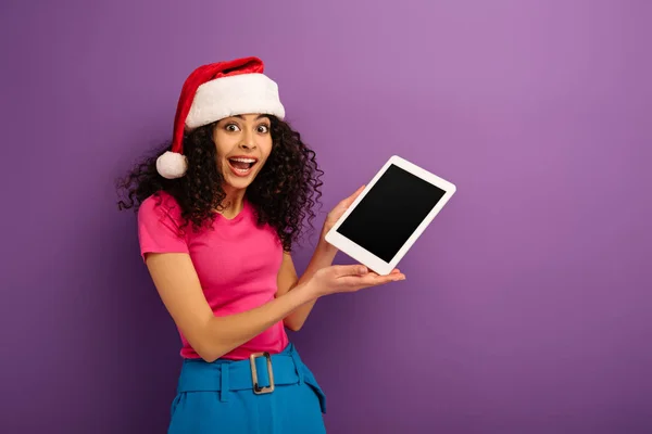 Aufgeregtes Mischlingsmädchen mit Weihnachtsmann-Hut zeigt digitales Tablet mit leerem Bildschirm auf lila Hintergrund — Stockfoto