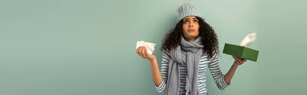 Tiro panorámico de niña bi-racial enferma en sombrero caliente y bufanda mirando hacia arriba mientras sostiene servilletas de papel sobre fondo gris - foto de stock