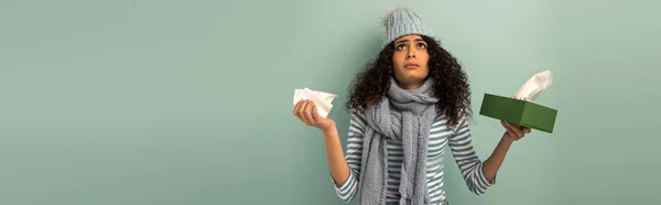 Panoramic shot of diseased bi-racial girl in warm hat and scarf looking up while holding paper napkins on grey background — Stock Photo