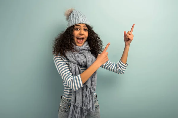 Sorprendida chica bi-racial en sombrero caliente y bufanda apuntando con los dedos mientras mira a la cámara sobre fondo gris - foto de stock