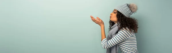 Side view of bi-racial girl in warm hat and scarf sending air kiss on grey background, panoramic shot — Stock Photo