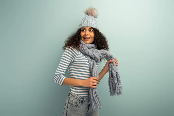 Щаслива бі-расова дівчина в теплому капелюсі і шарфі, дивлячись на сірий фон — стокове фото