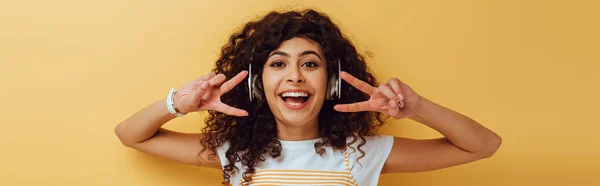 Panoramic shot of excited mixed race girl in wireless headphones showing victory gesture on yellow background — Stock Photo