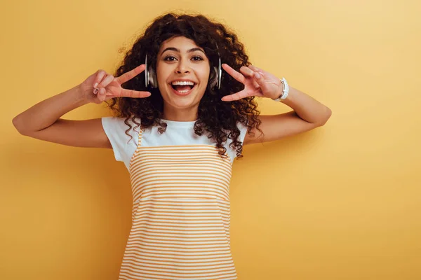 Cheerful mixed race girl in wireless headphones showing victory gesture on yellow background — Stock Photo