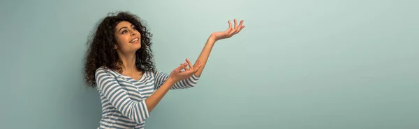 Panoramic shot of cheerful bi-racial girl looking up while standing with outstretched hands on grey background — Stock Photo