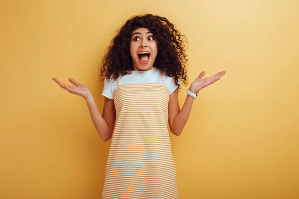 Shocked mixed race girl looking away while standing with open arms on yellow background — Stock Photo