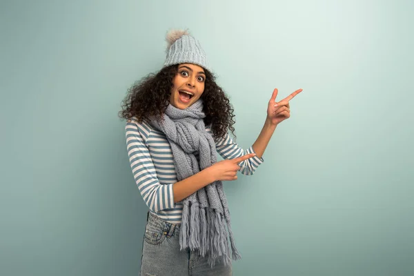 Потрясенная двурасовая девушка в теплой шляпе и шарфе, стоящая и смотрящая в камеру, указывая пальцами на сером фоне — стоковое фото