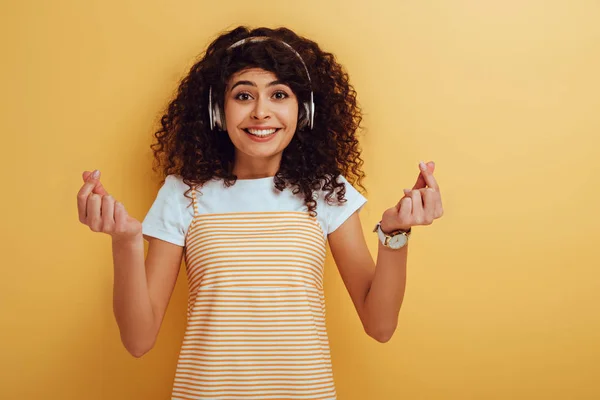 Excitada chica de raza mixta en auriculares inalámbricos que muestran el gesto ganador en el fondo amarillo - foto de stock