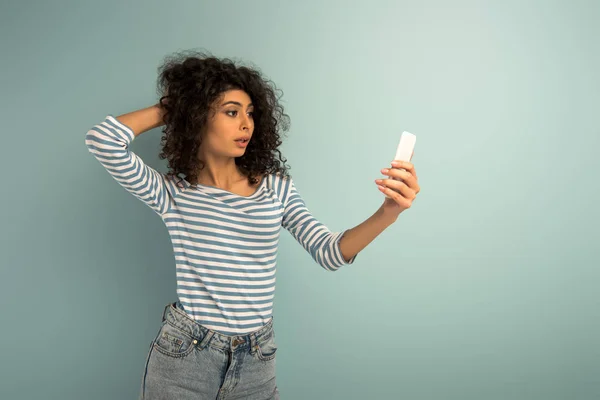 Atractiva chica bi-racial tocando el cabello mientras toma selfie en el teléfono inteligente sobre fondo gris - foto de stock