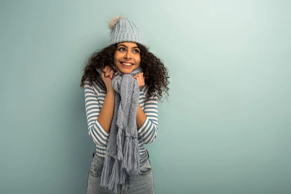 Симпатичная двурасовая девушка в теплой шляпе и шарфе улыбается и смотрит в сторону на сером фоне — стоковое фото
