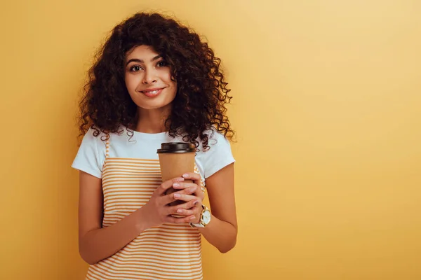 Jolie fille bi-raciale souriant à la caméra tout en tenant du café pour aller sur fond jaune — Photo de stock