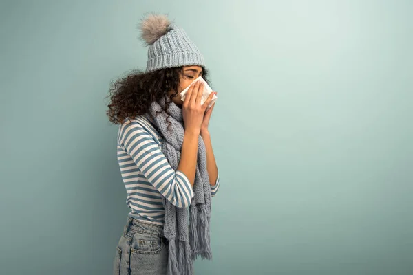 Diseased bi-racial girl in hat and scarf sneezing in napkin on grey background — Stock Photo