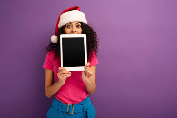 Cheerful mixed race girl in santa hat showing digital tablet with blank screen on purple background — Stock Photo