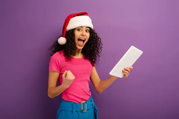 Excited mixed race girl in santa hat showing winner gesture while holding digital tablet on purple background — Stock Photo