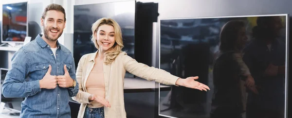 Colpo panoramico di fidanzato che mostra mi piace e sorridente ragazza che punta con mano a una nuova tv nel negozio di elettrodomestici — Foto stock