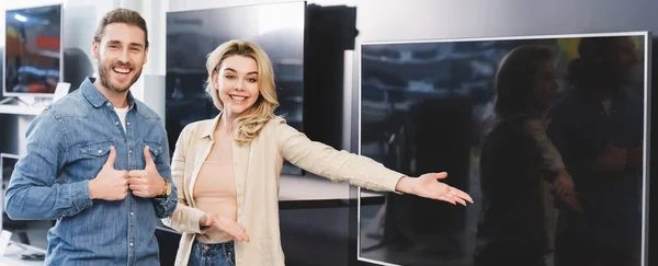 Plano panorámico de novio mostrando gustos y novia sonriente señalando con la mano en la nueva televisión en la tienda de electrodomésticos - foto de stock