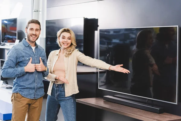 Namorado mostrando gostos e sorrindo namorada apontando com a mão para a nova tv na loja de eletrodomésticos — Fotografia de Stock