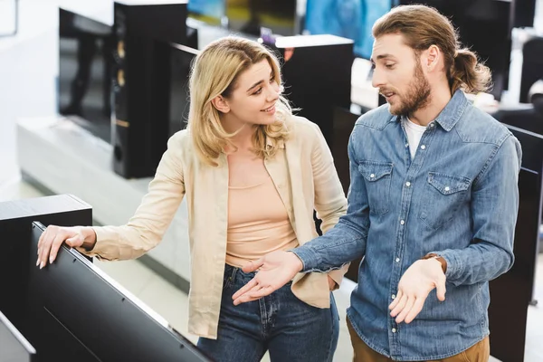 Boyfriend showing shrug gesture and talking with smiling girlfriend near new tv in home appliance store — Stock Photo