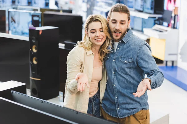 Novio y novia sonriente señalando con las manos en la nueva televisión en la tienda de electrodomésticos - foto de stock