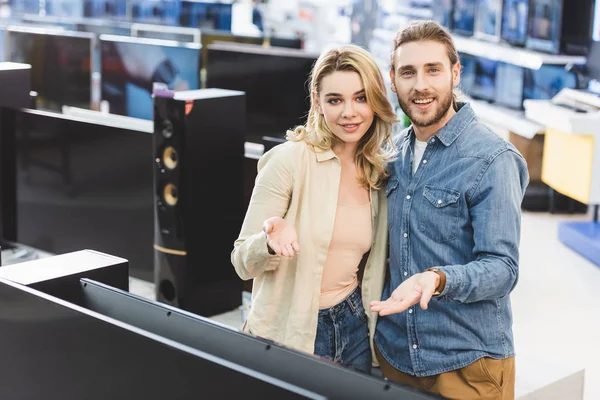 Novio y novia sonriente señalando con las manos en la nueva televisión en la tienda de electrodomésticos - foto de stock