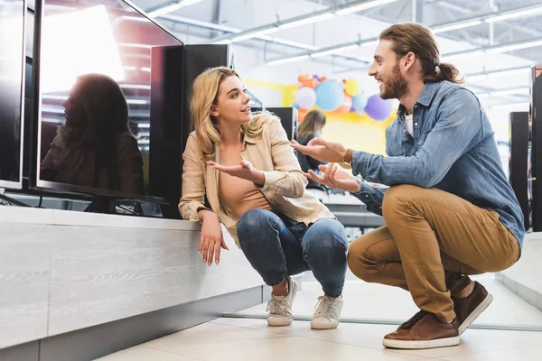 Lächelnder Freund und Freundin zeigt mit den Händen auf neuen Fernseher und redet im Haushaltsgerätegeschäft — Stockfoto