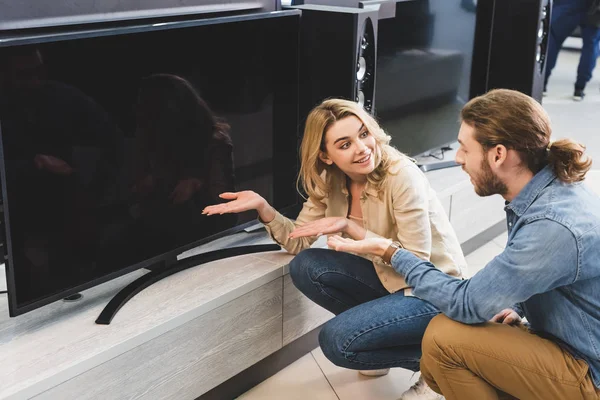 Freund und lächelnde Freundin zeigen mit den Händen auf neuen Fernseher und reden im Haushaltsgerätegeschäft — Stockfoto