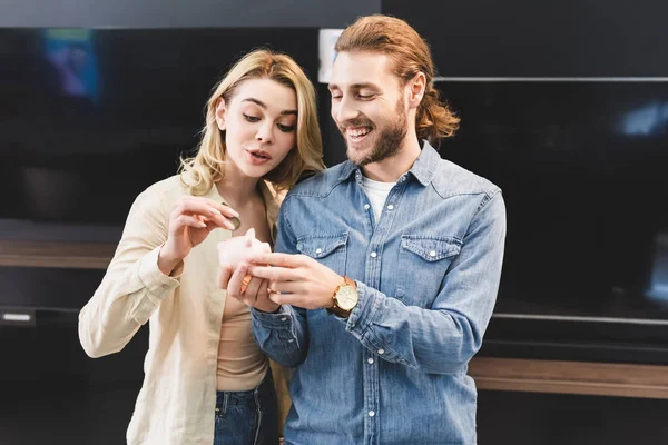 Smiling boyfriend holding piggy bank and girlfriend putting coin into it in home appliance store — Stock Photo