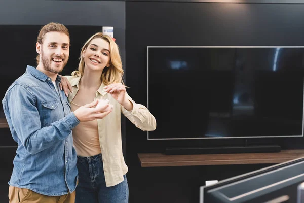 Smiling boyfriend holding piggy bank and girlfriend putting coin into it in home appliance store — Stock Photo