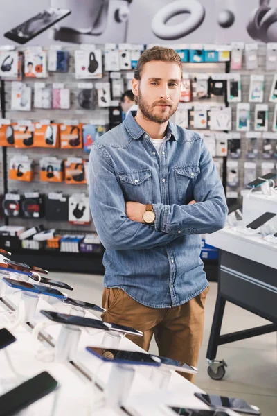 Schöner Mann mit verschränkten Armen steht neben neuen Smartphones im Haushaltsgerätegeschäft — Stockfoto