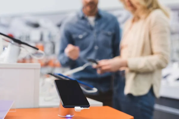 Selective focus of smartphone and boyfriend with girlfriend on background in home appliance store — Stock Photo