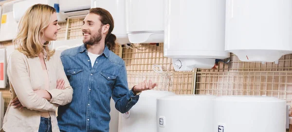 Panoramic shot of smiling boyfriend and girlfriend talking near boilers in home appliance store — Stock Photo