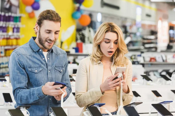 Shocked boyfriend and girlfriend looking at new smartphones in home appliance store — Stock Photo