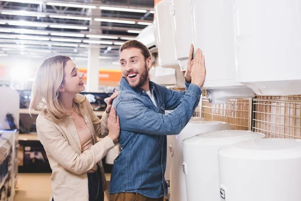 Petit ami souriant touchant chaudière et regardant petite amie dans le magasin d'appareils ménagers — Photo de stock