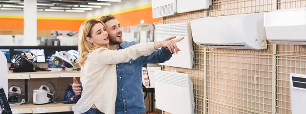 Panoramic shot of smiling boyfriend and girlfriend pointing with fingers at air conditioner in home appliance store — Stock Photo