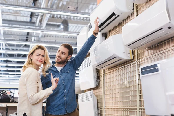 Boyfriend pointing with hand at air conditioner and girlfriend showing no gesture in home appliance store — Stock Photo