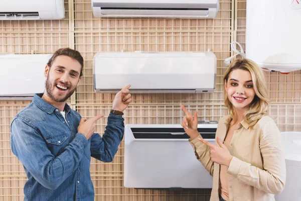 Novio sonriente y novia señalando con los dedos en el aire acondicionado en la tienda de electrodomésticos - foto de stock