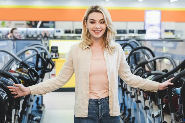Donna sorridente e attraente in piedi vicino a nuovi aspirapolvere nel negozio di elettrodomestici — Foto stock