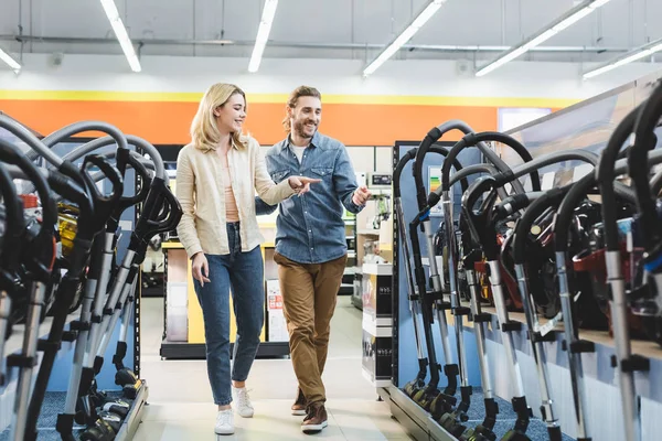 Novio y novia sonrientes apuntando con los dedos a las aspiradoras en la tienda de electrodomésticos - foto de stock