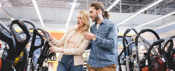 Panoramic shot of smiling boyfriend pointing with finger and looking at vacuum cleaner with shocked girlfriend in home appliance store — Stock Photo